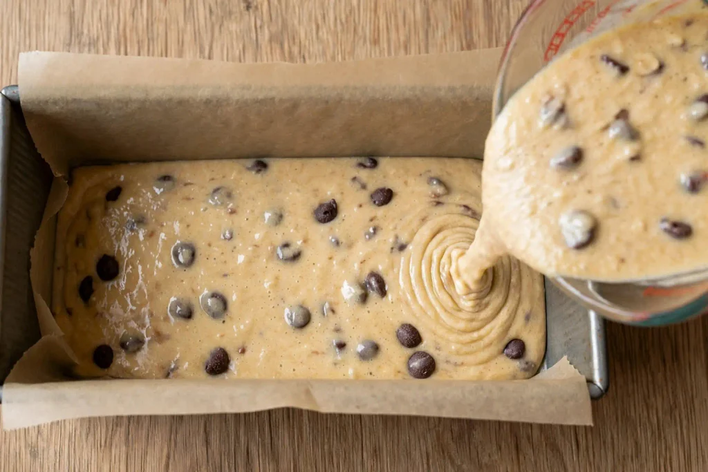 Chocolate Chip Banana Bread 3 Banana bread batter being poured into a lined loaf pan with chocolate chips visible